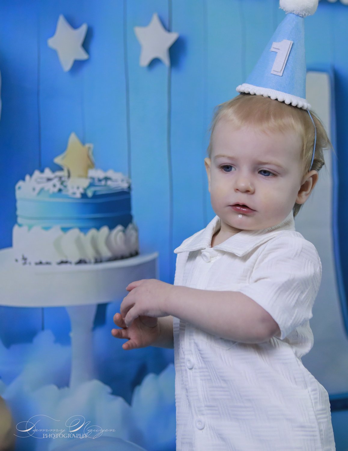 A one-year-old in a party hat standing beside a blue birthday cake.