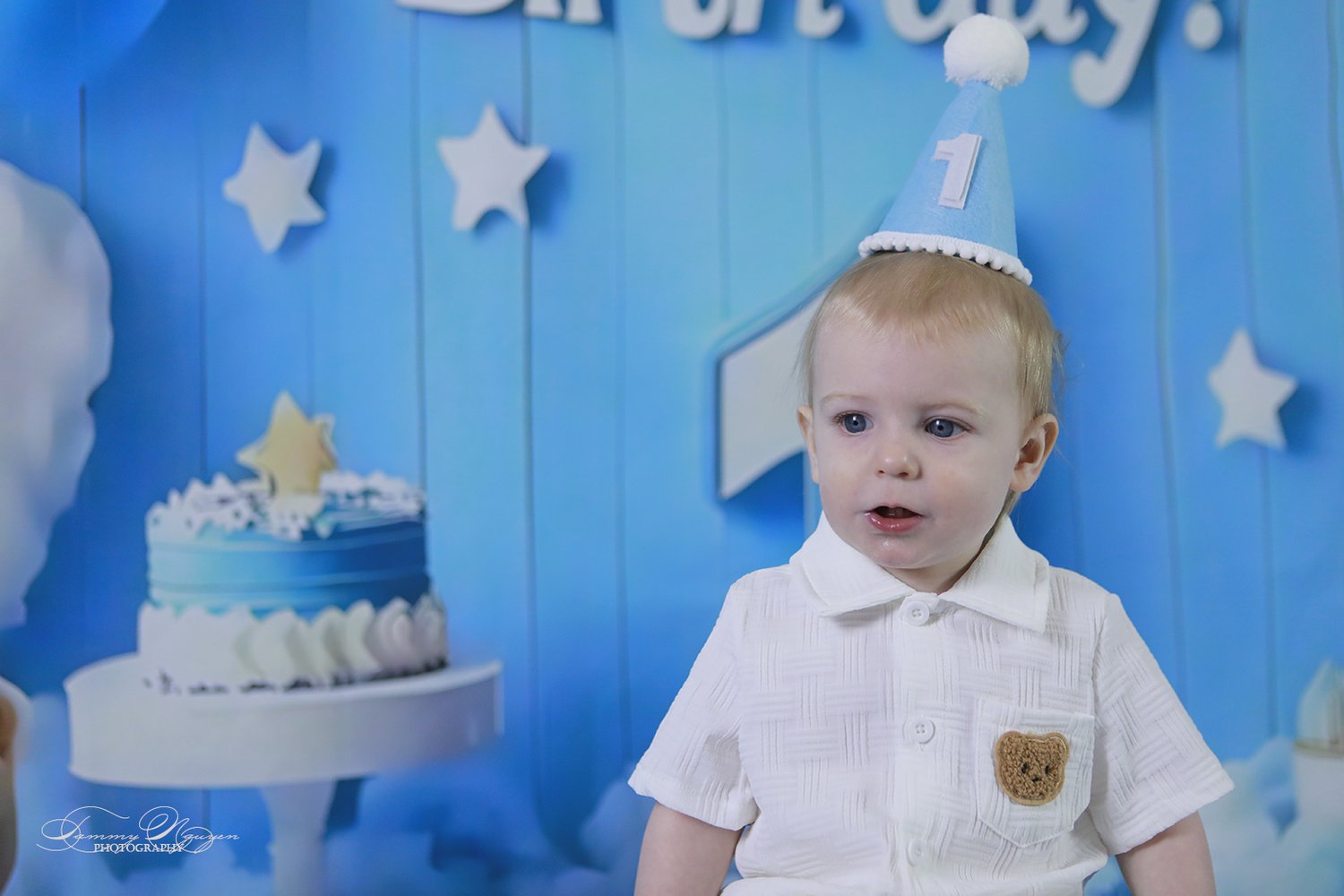 A one-year-old in a party hat beside his birthday cake.