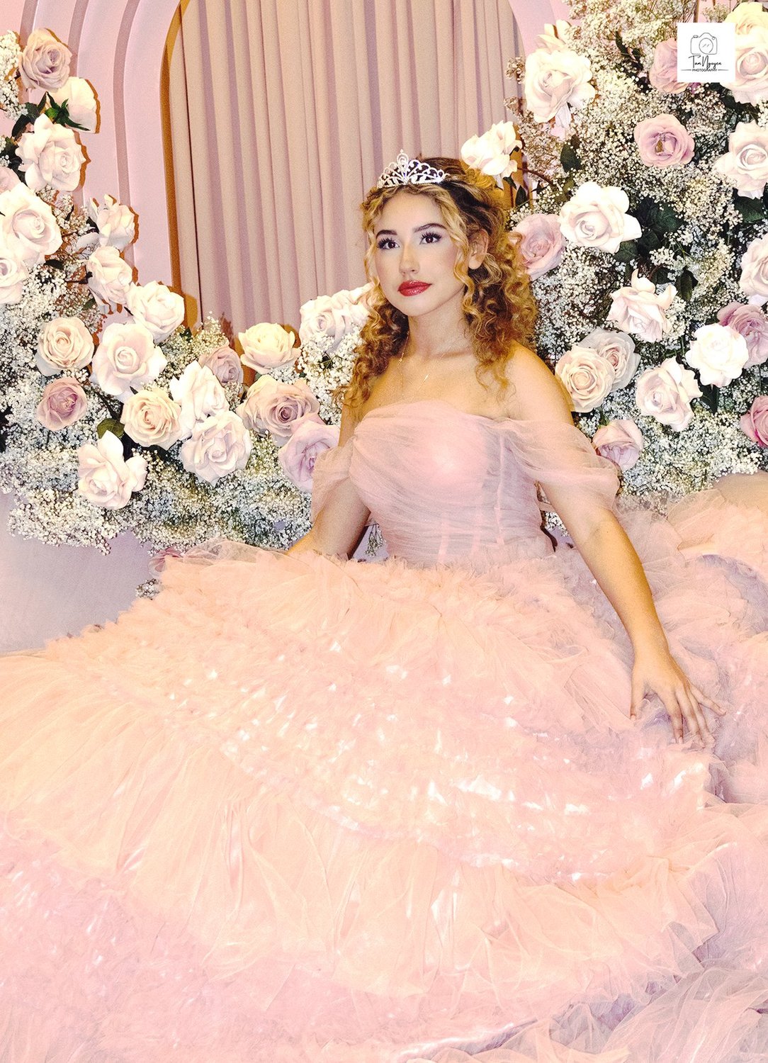 A young woman in a quinceanera gown in front of a wall of roses.